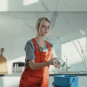 A woman wearing orange overalls cleans a kitchen counter with a rag and pail in a modern kitchen.